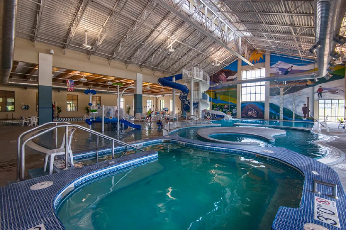 A vibrant indoor water park scene with swimming pools, colorful water slides, and playful murals. Lounge chairs line the pool, and children are seen enjoying the amenities.