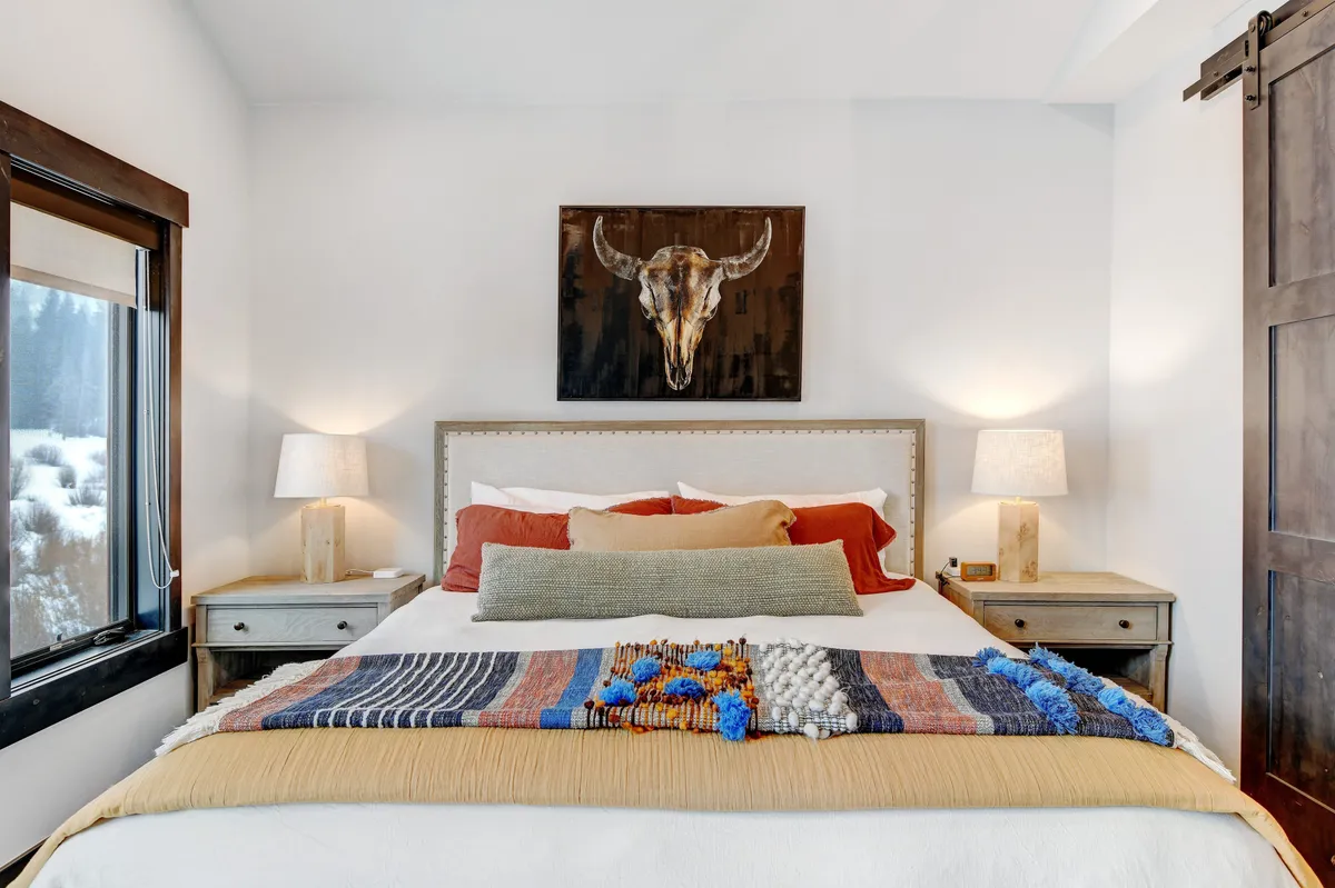 A cozy bedroom with a bed adorned with decorative throw pillows, mounted bull skull artwork above the headboard, and matching nightstands with warm lamps on each side. The serene lighting and soft linens create a tranquil, welcoming atmosphere.