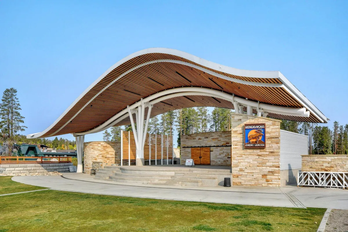 An exterior shot of the Rendezvous Event Center, featuring its unique wave-like roof structure, stone walls, and an open stage area ready for concerts or community events. The presence of event signage and seating underscores its importance as a hub for local culture and entertainment.