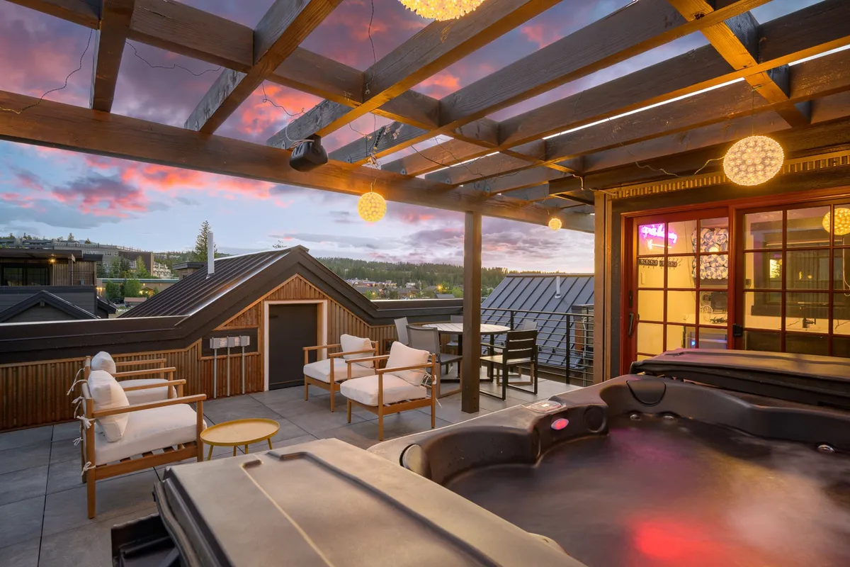 An inviting rooftop patio at sunset featuring a luxurious private hot tub, plush outdoor seating beneath a stylish pergola adorned with hanging lights, and a serene view of a glowing sky. The space is primed for both relaxation and social gatherings.