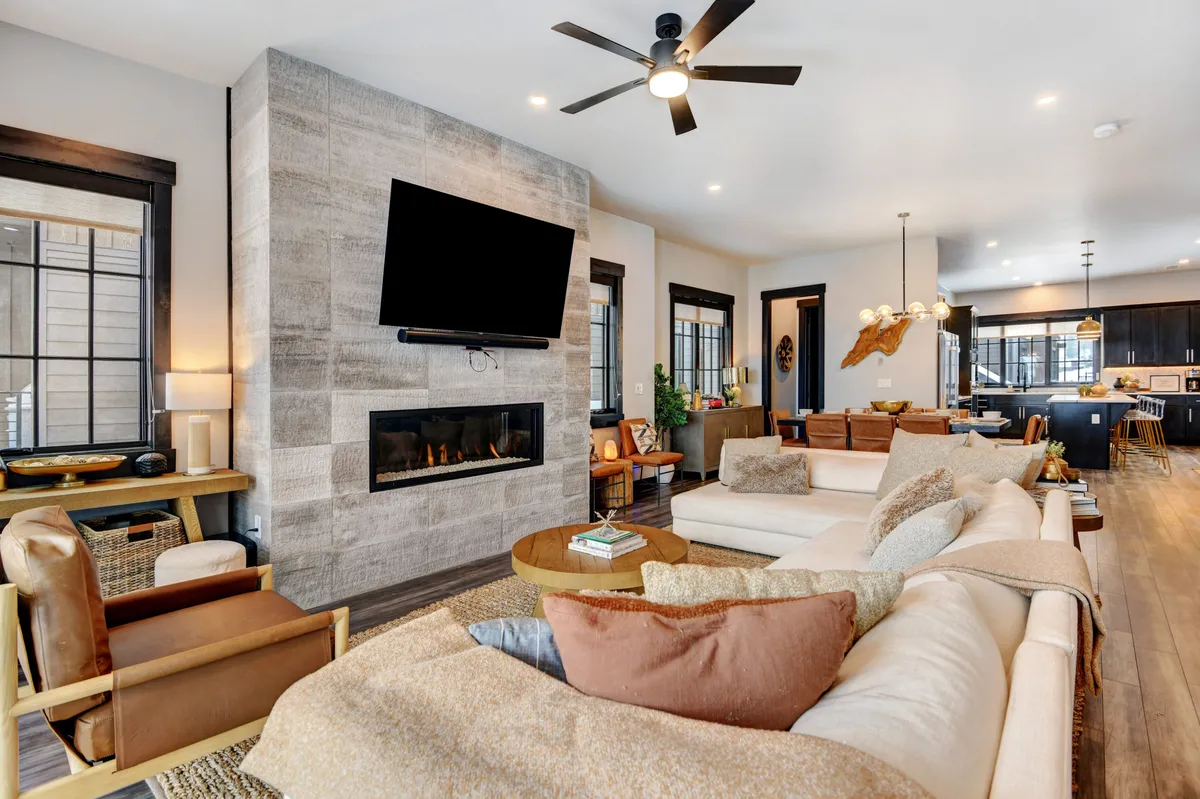 A spacious open-concept living room featuring a striking doublewide fireplace as the focal point, with a large plush sofa, wall-mounted television, and an adjacent dining area. Warm lighting and cozy decor make the space inviting for gatherings and relaxation.