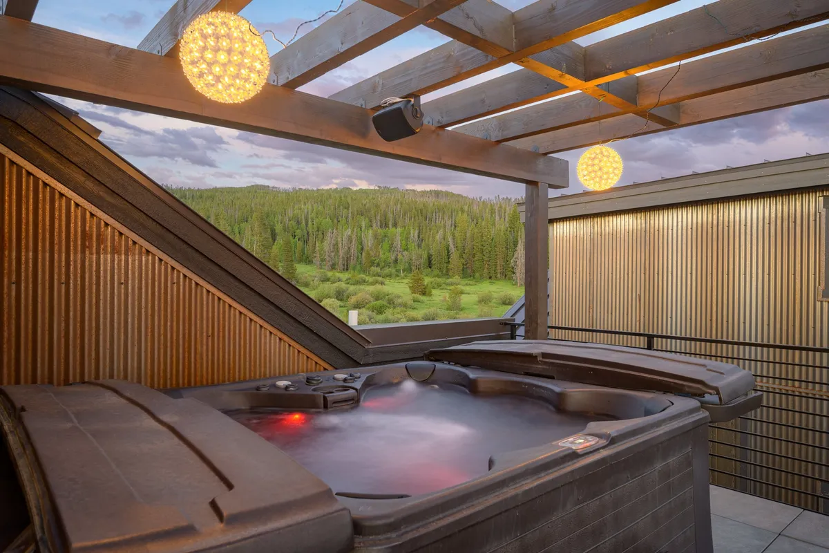 A tranquil rooftop hot tub surrounded by lush greenery, strung with warm hanging lights. The scene overlooks natural vistas, inviting guests to soak in the peaceful ambiance at sunset or under the stars.