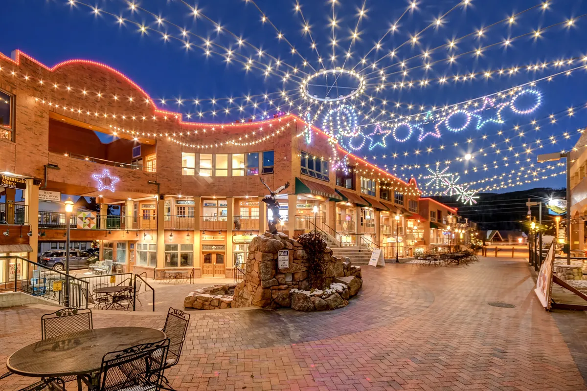 An exterior view of a brick pathway flanked by building facades, strung with glowing lights and accented by decorative sculptures. The scene exudes warmth, joy, and the energy of al fresco dining or community gatherings, perfect for winter or summer evenings.