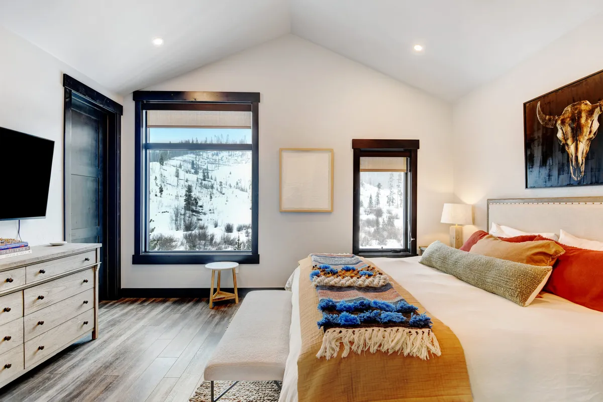 A tranquil bedroom with large windows framing a scenic mountain view, a comfortable bed with a decorative throw, and a wooden dresser topped with a TV. The natural light and soothing colors enhance the sense of calm and retreat.